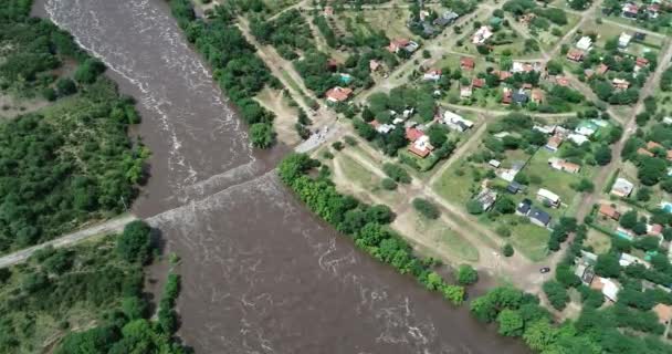 Escena senital aérea de amplio río fuerte que fluye y crece hacia la ciudad rural, que fluye sobre el puente. Descendig mostrando detalle de la construcción. Flood Mina clavero, traslasierra, Córdoba, Argentina — Vídeos de Stock