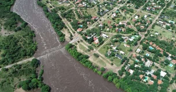 Senitalna scena dronów lotniczych o szerokim brązowym strumieniu rzeki płynącej i rosnącej w kierunku wiejskiego miasta, przepływającej przez most. Powódź w Mina clavero, traslasierra, Cordoba, argentina — Wideo stockowe