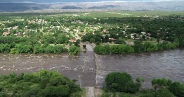 Kahverengi güçlü nehrin köprüden geçtiği hava aracı sahnesi. Mina Clavero 'da sel. Şehrin sokaklarında büyüyen nehir. İnşaata doğru uçarken, arka planda evler. Cordoba, Arjantin