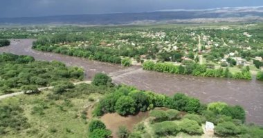 Kahverengi güçlü nehrin köprüden geçtiği hava aracı sahnesi. Mina Clavero 'da sel. Şehrin sokaklarında büyüyen nehir. İnşaata doğru uçarken, arka planda evler. Cordoba, Arjantin