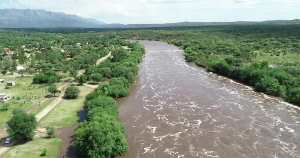 Scena dronów lotniczych z brązową, silną rzeką przelatującą przez most. Powódź w Mina clavero. Rosnąca rzeka nad ulicami miasta. Latanie nad wodą w kierunku budowy. Kordoba, Argentyna — Wideo stockowe