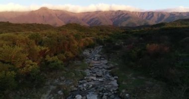 Ağaçlık araziler arasındaki kayalık dağ nehrinin üzerinde gün batımının hava aracı görüntüsü. Arka planda boylamsal bulut olan dağ zinciri. San Javier, Cordoba, Arjantin 'de altın saat
