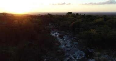 Ağaçlık araziler arasındaki kayalık dağ nehrinin üzerinde turuncu günbatımının hava aracı görüntüsü. Arka planda dağlar ve güneş. San Javier, Cordoba, Arjantin 'de altın saat