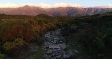 Ağaçlık araziler arasındaki kayalık dağ nehrinin üzerinde gün batımının hava aracı görüntüsü. Arka planda boylamsal bulut olan dağ zinciri. San Javier, Cordoba, Arjantin 'de altın saat