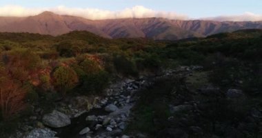 Ağaçlık araziler arasındaki kayalık dağ nehrinin üzerinde gün batımının hava aracı görüntüsü. Arka planda boylamsal bulut olan dağ zinciri. San Javier, Cordoba, Arjantin 'de altın saat
