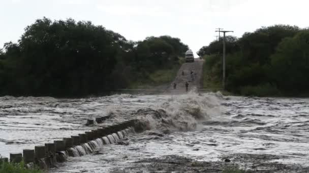 Slow motion of strong river passing over bridge. Turbulent violent water flow over construction. People and fence across river at background. Flood in Mina Clavero, growing river, Cordoba, Argentina — Stockvideo