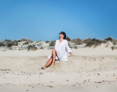 Beyaz elbiseli genç bir kadın sahilde oturuyor. Seyahat ve tatil kavramı.