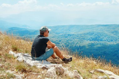 Bir tepenin tepesinde dinlenen turist güneşli bir günde dağların muhteşem manzarasını seyrediyor.
