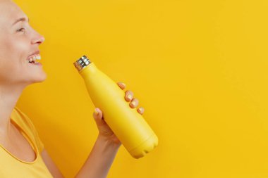 Sarı şişeden içen kadın sarı arka planda ve gülümsüyor.