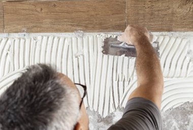 Zanaatkar fayans yapıştırıcısını fayanssız bir evin zemin yüzeyine serpiştirir, eller üst görüntüyü kapatır. Yeni bina inşaatı veya tadilatı. Porselen taş savaşıyla döşeme