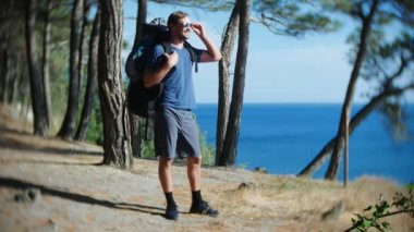 yakışıklı turist adam büyük bir sırt çantası ile bir çam koru ile dik sarp deniz kıyı kenar gitmektedir. deniz manzara muhteşem bir görünüm ile. 4k.