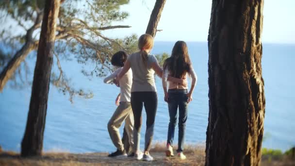 Una madre feliz familia y su hijo y su hija van al borde de una costa empinada con un pinar. disfrutan de una magnífica vista del paisaje marino. 4k . — Vídeos de Stock