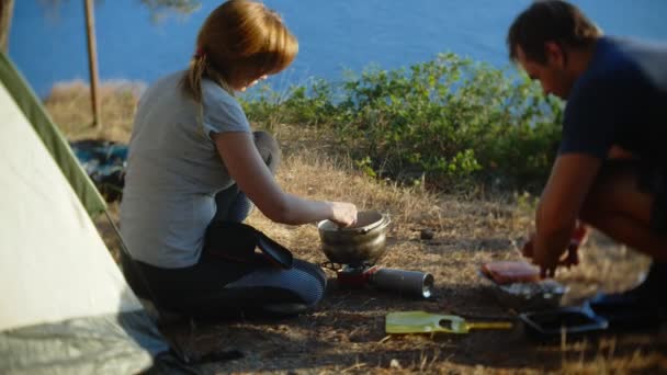 Un hombre y una mujer, los campistas, cocina comida al lado de una tienda de campaña en el borde de una costa empinada en un pinar con una magnífica vista del paisaje marino. 4k — Vídeo de stock