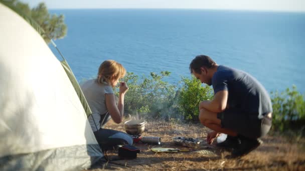 Un uomo e una donna, i campeggiatori, cucinano accanto a una tenda sul bordo di una ripida costa in una pineta con una magnifica vista sul paesaggio marino. 4k — Video Stock