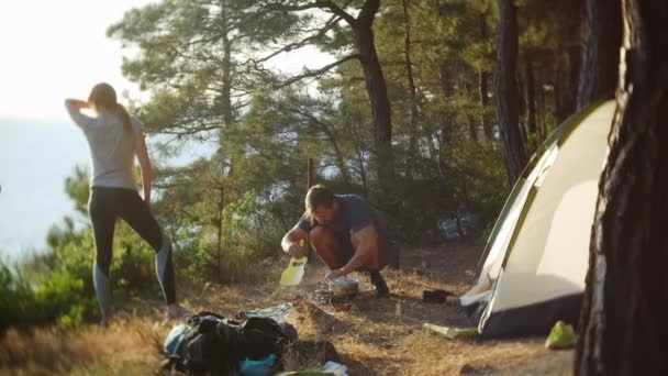 Un hombre y una mujer, los campistas, cocina comida al lado de una tienda de campaña en el borde de una costa empinada en un pinar con una magnífica vista del paisaje marino. 4k — Vídeos de Stock