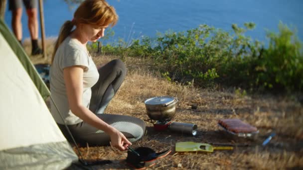 Un hombre y una mujer, los campistas, cocina comida al lado de una tienda de campaña en el borde de una costa empinada en un pinar con una magnífica vista del paisaje marino. 4k — Vídeo de stock