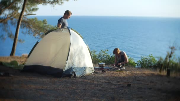 Un homme et une femme, les campeurs, cuisinent à côté d'une tente sur le bord d'une côte escarpée dans une pinède avec une vue magnifique sur le paysage marin. 4k — Video