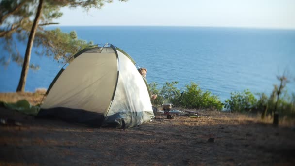 Eine Frau, die Camperin, kocht neben einem Zelt am Rande einer steilen Küste in einem Kiefernhain mit herrlichem Blick auf die Meereslandschaft. 4k. — Stockvideo