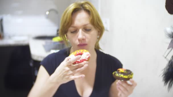 Γυναίκα τρώει γλυκά ντόνατς με γλάσο χρωματιστό στην κουζίνα στο σπίτι και χρησιμοποιώντας το smartphone της, 4k, Θάμπωμα φόντου — Αρχείο Βίντεο