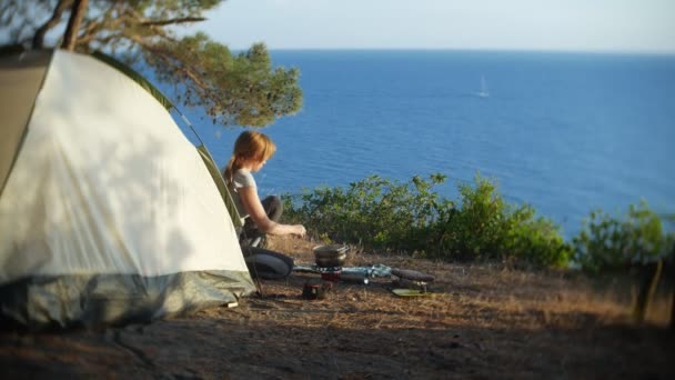 Eine Frau, die Camper, kocht neben einem Zelt am Rande einer Steilküste in einem Kiefernhain mit herrlichem Blick auf die Meereslandschaft. im Hintergrund schwimmt eine Jacht am Meer, und eine — Stockvideo