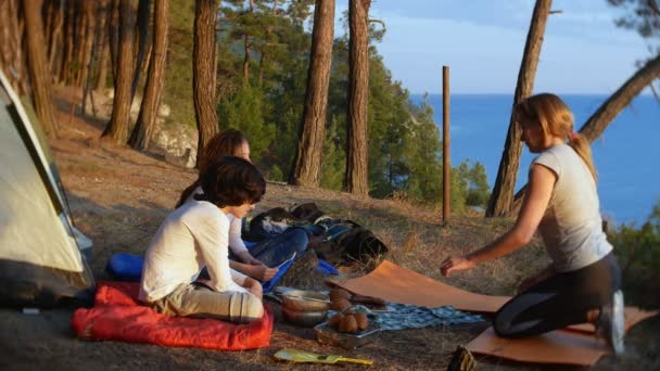 Una familia feliz de turistas, comiendo comida de campamento, junto a una tienda de campaña en el borde de una costa empinada en un pinar con una magnífica vista del paisaje marino. 4k . — Vídeo de stock