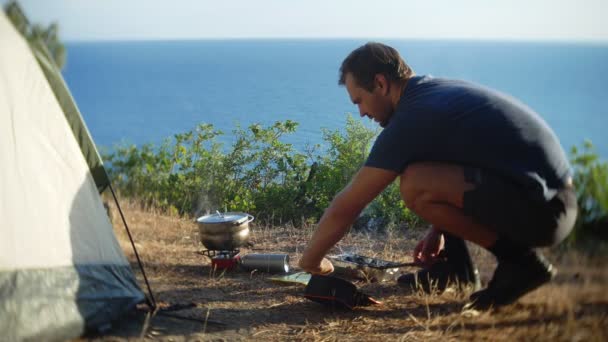 Un hombre los campistas, cocina comida al lado de una tienda de campaña en el borde de una costa empinada en un pinar con una magnífica vista del paisaje marino. 4k — Vídeos de Stock