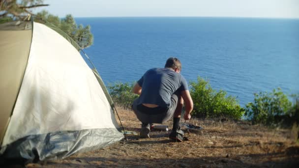 En man campare, kockar mat bredvid ett tält med en magnifik utsikt över havet, i utkanten av en brant kustlinje i en pinjelund landskap. 4k — Stockvideo