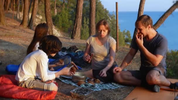 Una familia feliz de turistas, comiendo comida de campamento, junto a una tienda de campaña en el borde de una costa empinada en un pinar con una magnífica vista del paisaje marino. 4k . — Vídeos de Stock