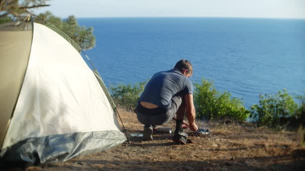 Egy ember a táborozók, szakácsok élelmiszer egy sátor, egy meredek tengerparton, egy fenyőligetben szélén fekszik, csodálatos kilátással a tenger mellett fekvő. 4k — Stock videók