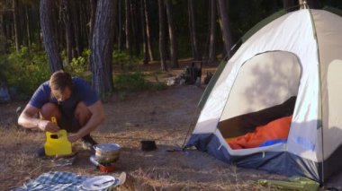 bir adam kampçılar, aşçılar yemek denizin muhteşem manzaralı pine Grove dik bir sahil kenarında bir çadır yanındaki manzara. 4k