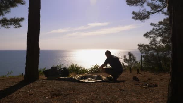 Een mannelijke toeristische regelt een tent aan de rand van een steile kust kust in een pijnbomenbos met een prachtig uitzicht op het Zeegezicht. 4k. — Stockvideo