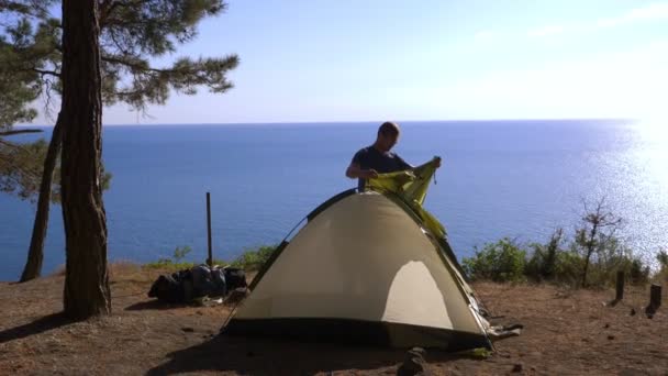 Мужчина-турист ставит палатку на краю крутого прибрежного берега в сосновой роще с великолепным видом на море. 4k . — стоковое видео