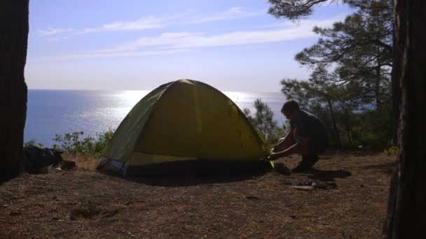 Um turista masculino organiza uma barraca na borda de uma costa costeira íngreme em um bosque de pinheiros com uma vista magnífica da paisagem marinha. 4k . — Vídeo de Stock