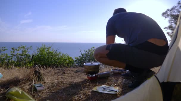 Un hombre los campistas, cocina comida al lado de una tienda de campaña en el borde de una costa empinada en un pinar con una magnífica vista del paisaje marino. 4k — Vídeo de stock