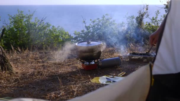 Un hombre los campistas, cocina comida al lado de una tienda de campaña en el borde de una costa empinada en un pinar con una magnífica vista del paisaje marino. 4k — Vídeo de stock