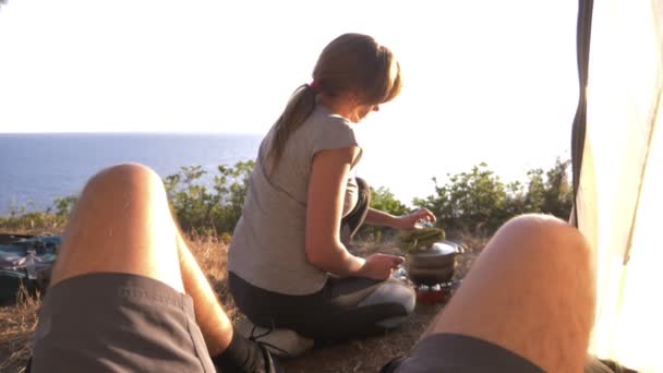 Um homem e uma mulher, os campistas, cozinham comida ao lado de uma tenda na beira de um litoral íngreme em um pinheiro com uma vista magnífica da paisagem do mar. 4k — Vídeo de Stock