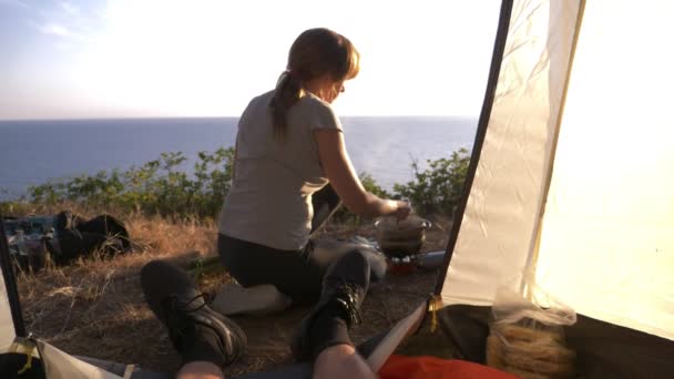 Un hombre y una mujer, los campistas, cocina comida al lado de una tienda de campaña en el borde de una costa empinada en un pinar con una magnífica vista del paisaje marino. 4k — Vídeos de Stock