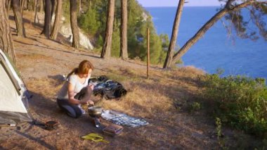 bir kadın, kampçılar, yanında bir çadır deniz manzara muhteşem manzaralı pine Grove dik bir sahil kenarında yemek pişiriyor. 4k
