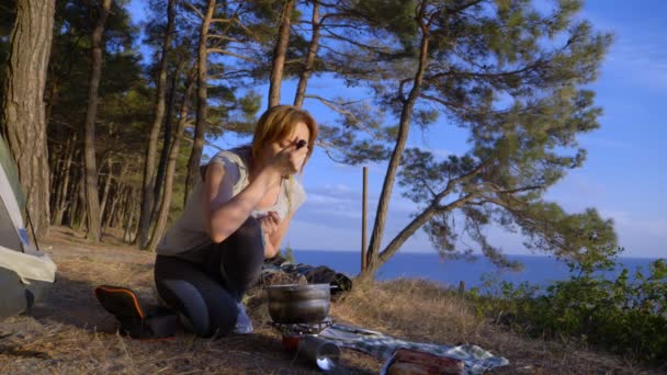 Женщина, отдыхающие, готовит еду рядом с палаткой на краю крутой береговой линии в сосновой роще с великолепным видом на морской пейзаж. 4k — стоковое видео