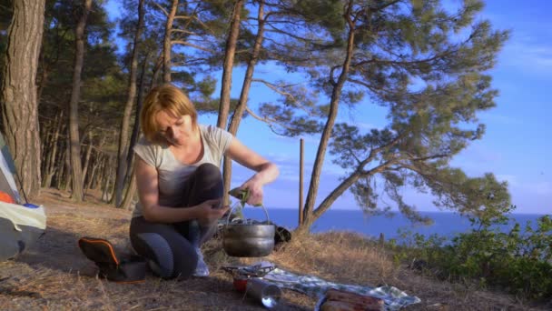 Женщина, отдыхающие, готовит еду рядом с палаткой на краю крутой береговой линии в сосновой роще с великолепным видом на морской пейзаж. 4k — стоковое видео