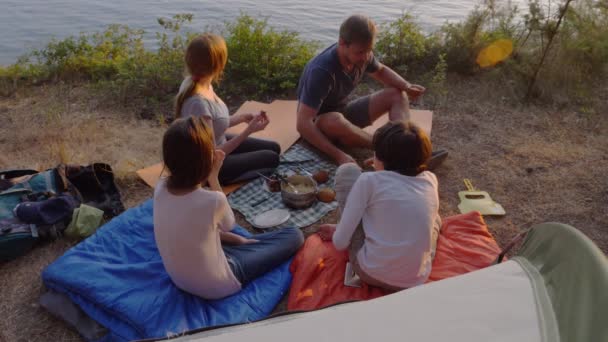 Una familia feliz de turistas, comiendo comida de campamento, junto a una tienda de campaña en el borde de una costa empinada en un pinar con una magnífica vista del paisaje marino. 4k . — Vídeos de Stock