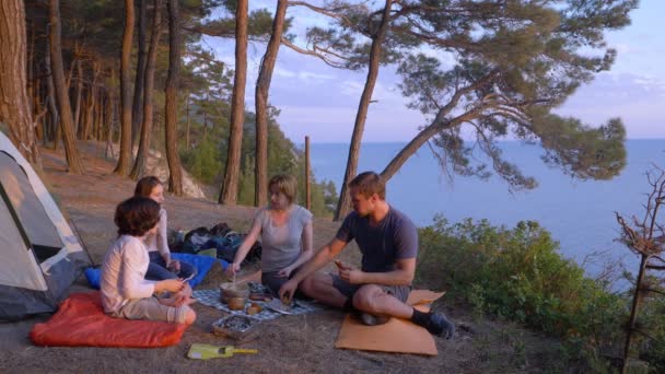 Счастливая семья туристов, питающихся кемпингом, рядом с палаткой на краю крутой береговой линии в сосновой роще с великолепным видом на море. 4k . — стоковое видео