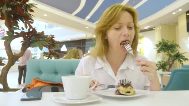 Una mujer con una camisa blanca come con un cuchillo y tenedor un bollo de canela en un café y bebe café. Concepto de desayuno. 4k — Vídeos de Stock