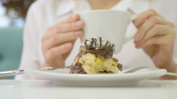 Жінка в білій сорочці їсть з ножем і виделкою булочку кориці в кафе і п'є каву. Концепція сніданку. 4k — стокове відео