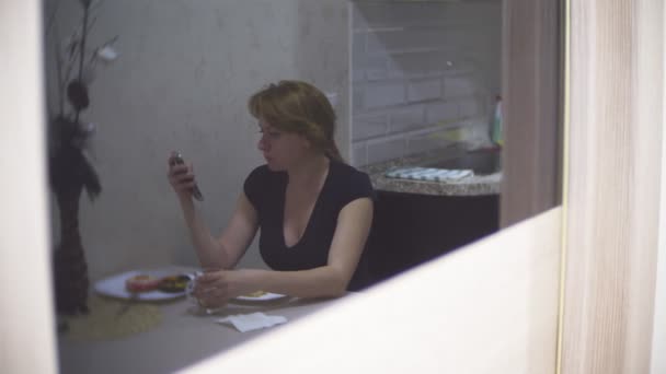 Женщина ест сладкие пончики с цветной глазурью на кухне дома и с помощью своего смартфона, 4k, размытый фон — стоковое видео