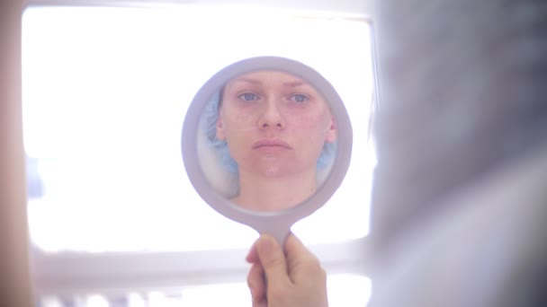 La femme regarde son visage dans le miroir après la procédure dans le cabinet des médecins de l'esthéticienne. 4k . — Video