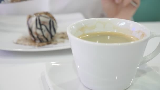 Uma mulher de camisa branca come com uma faca e garfo um rolo de canela de pão em um café e bebe café. Conceito de pequeno almoço. 4k — Vídeo de Stock