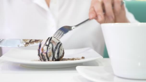 En kvinna i en vit skjorta äter med kniv och gaffel en bulle kanelbulle i ett café och drycker kaffe. Frukost koncept. 4k — Stockvideo