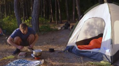 bir adam kampçılar, aşçılar yemek denizin muhteşem manzaralı pine Grove dik bir sahil kenarında bir çadır yanındaki manzara. 4k