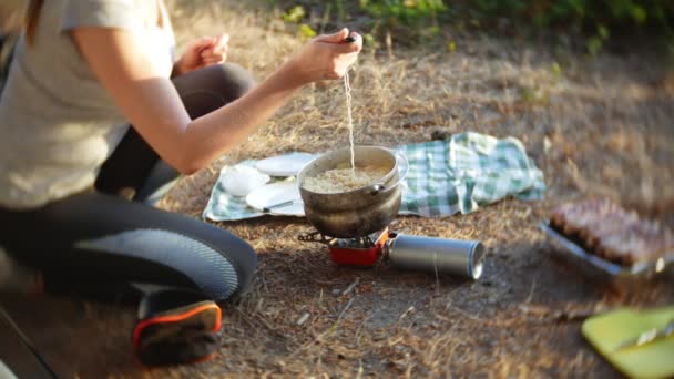 Женщина, отдыхая, готовит еду рядом с палаткой на краю крутой береговой линии в сосновой роще с великолепным видом на море. 4k . — стоковое видео